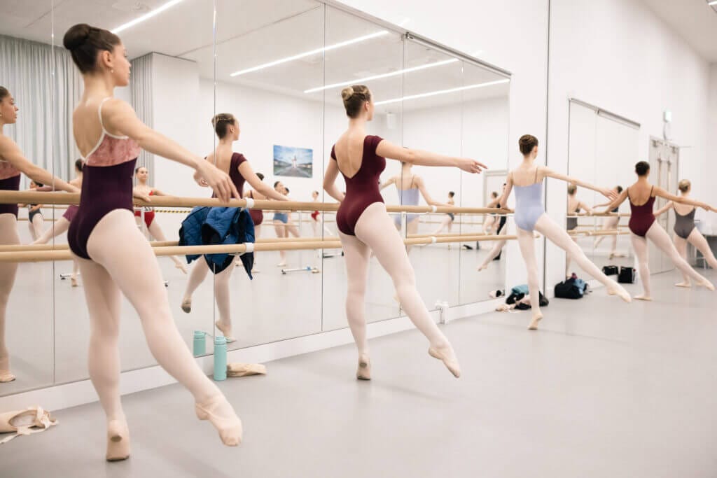 Image showing candidates in warm up class at the barre Female dancers in tights leotards and ballet shoes with hair in a bun rise up on a demi with leg a la seconde